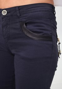 Pantalones de mezclilla azul marino con un bolsillo lateral negro texturizado con detalles perforados, cierre con botón de metal y un diseño ajustado.