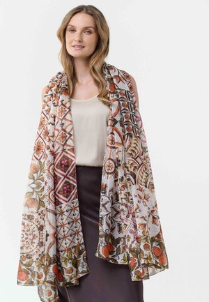 Frau trägt ein cremefarbenes ärmelloses Oberteil und einen dunklen Rock, bedeckt mit einem großen, gemusterten Schal in Herbstfarben.