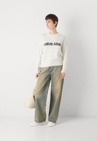 Hvit strikket hettegenser med svart "calvin klein" logo, parret med høy midje, vide, falmede blå jeans og hvite sneakers, holder en beige veske.