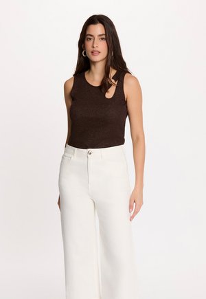 Vrouw met lang donker haar die een mouwloos donkerbruin topje met uitsnijding en witte, hooggetailleerde, wijde broek draagt.