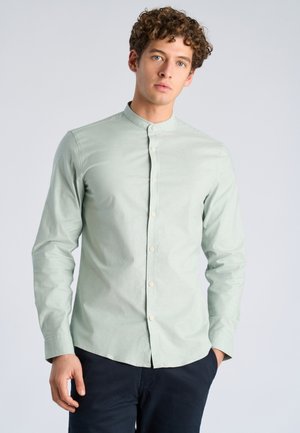 Junger Mann mit lockigem Haar, der ein hellgrünes Hemd mit Mandarin-Kragen und dunkle Hose trägt, steht vor einem schlichten hellen Hintergrund.