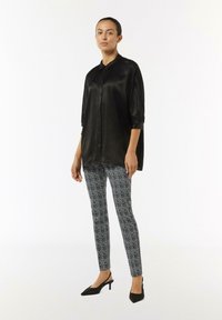 Schwarzes Satin-Hemd mit Knöpfen, 3/4-Ärmel, kombiniert mit figurbetonten schwarz-weiß gemusterten Hosen. Das Model trägt schwarze Absatzschuhe.