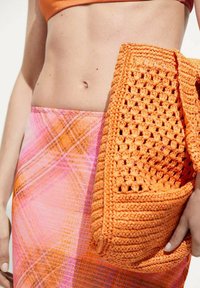 Sac en crochet orange avec des détails texturés, maintenu contre une jupe en plaid rose et orange en tissu léger.