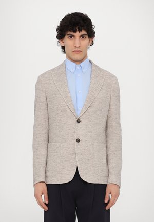 Blazer chiaro e strutturato con chiusura a un pulsante, due tasche frontali e rever a giorno, indossato sopra una camicia azzurro chiaro.