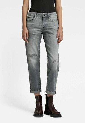 Personne portant un jean gris délavé et usé, retroussé aux chevilles, avec des bottines en cuir marron foncé et un haut noir sans manches.