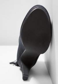 Botte à talon haut en daim noir à bout pointu avec une semelle lisse, un bord scintillant et un détail à glands, reposant contre un sol et un mur blancs.