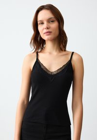 Zwarte camisole van ribbelstof, met kantafwerking langs de V-halslijn en verstelbare dunne banden. Zachte textuur en getailleerd ontwerp.
