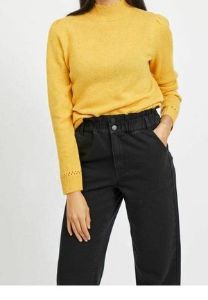 Kvinde iført en sennepsgul langærmet sweater med bølget udskæring og sorte højtaljede paperbag-bukser, med den ene hånd i lommen.