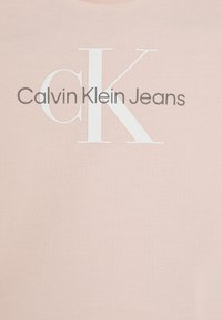 Vaaleanpunainen puuvillainen T-paita, jossa on suuri valkoinen "CK"-logo ja harmaalla "Calvin Klein Jeans" -teksti. Sileä pinta, lyhyet hihat, pyöreä pääntie.