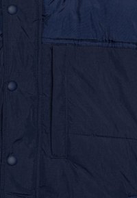 Materijal tamnoplave puf jakne s glatkom teksturom, koja ima zatvaranje na dugmad i vidljivu preklopnu džepu na prednjoj strani.