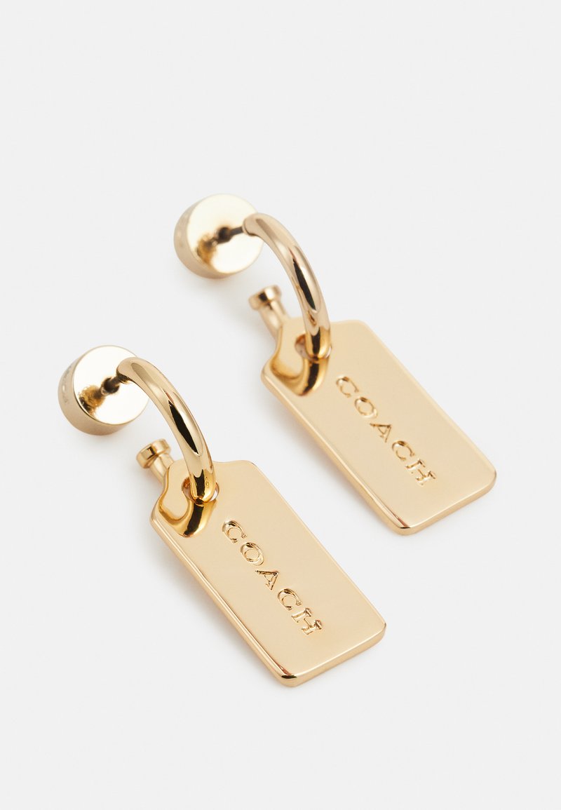 Des boucles d'oreilles en or présentent un design en forme de boucle avec des accents en forme de tag rectangulaire portant le nom de la marque. La surface est brillante et possède une texture lisse.