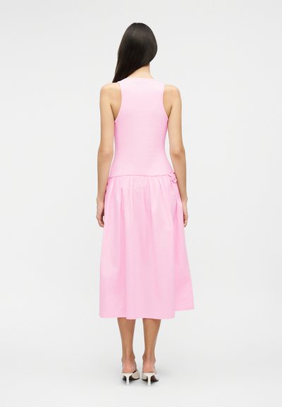 Ženska z dolgimi temnimi lasmi, oblečena v roza midi obleko brez rokavov in bele visoke pete, stoji z obrazom obrnjena proč na enobarvnem ozadju.