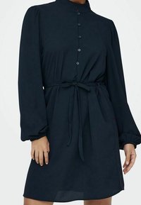 Mörkblå klänning med hög krage, långa puffärmar, knappstängning framtill och knytmidja. Tillverkad av ett lent tyg med en avslappnad passform.