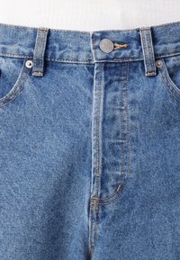 Jeans de denim azul claro com uma cintura alta, apresentando costura amarela em contraste, fecho de botão metálico e bolsos frontais.