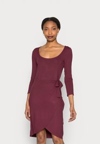 Vestido justo em malva com decote redondo, mangas 3/4 e detalhe de amarração lateral. O tecido tem uma textura suave e um design envolvente até ao joelho.
