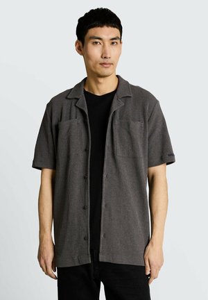 Uomo con camicia grigio scuro a maniche corte abbottonata sopra una T-shirt nera, su sfondo chiaro e uniforme.