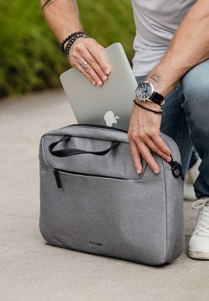 Šedá textilní taška na notebook se zipovým uzavřením a přední kapsou, uvnitř které je MacBook. Má dvojité rukojeti a hladkou strukturu.
