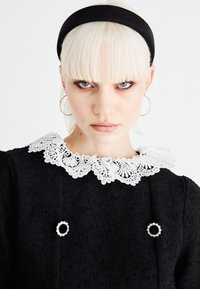 Črna teksturirana obleka z belim čipkastim ovratnikom in okrasnimi bisernimi detajli. Manekenka z črnim trakcem za lase in platinsko blond lasmi.