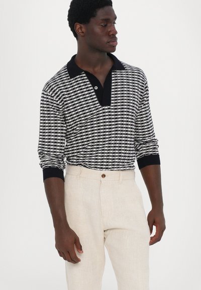 Black and white patterned long-sleeve polo shirt with a dark collar, paired with beige linen trousers featuring a button closure.