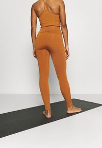 Pomarańczowy zestaw odzieży sportowej o teksturowanym wykończeniu, z dopasowaną bluzką i legginsami o wysokim stanie, prezentowany na osobie stojącej na macie do jogi.