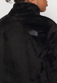 Persona che indossa una giacca nera pelosa con colletto alto e il logo "The North Face" sulla parte superiore della schiena, mostrando parte del suo profilo e un orecchino a cerchio.