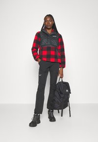 Rød og svart rutete fleece-pullover med et svart quiltet panel, parret med svarte bukser og en svart ryggsekk. Svarte sko synlige.