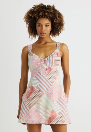 Femme aux cheveux bouclés portant une robe d'été sans manches à rayures multicolores avec un motif patchwork, mains dans les poches, fond neutre.