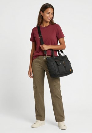 Frau trägt ein burgunderfarbenes T-Shirt und olivfarbene Cargohosen, trägt eine schwarze Tragetasche mit Schulterriemen über der rechten Schulter.