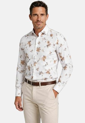 Man draagt een wit overhemd met lange mouwen met beige bloemenpatroon, beige broek en een bruine riem, staat met één hand in de zak.