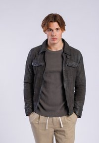 Chaqueta de cuero gris oscuro con dos bolsillos en el pecho, cuello redondo y detalles de costura visibles; superpuesta sobre una blusa texturizada de color verde oliva.