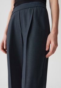Donkergrijze op maat gemaakte broek met een zachte textuur, voorzien van een ontspannen tailleband, zijplooien en schuine zakken. Close-up van de stofdetails.