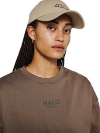 Młoda osoba z zaplecionymi włosami nosi beżową czapkę z napisem "OFF DUTY GEAR" oraz brązową bluzę z nadrukiem "HALO" z przodu.