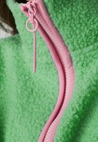 Giacca in pile verde con colletto alto, caratterizzata da una zip e un'esca pink a contrasto. Il tessuto ha un aspetto morbido e texture.