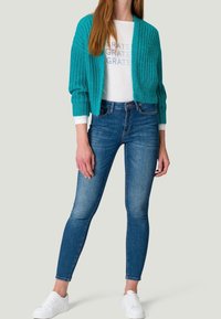 Türkiser Strickcardigan mit gerippter Textur, weißes Langarmshirt mit "GRATE"-Druck und dunkelblaue Skinny-Jeans, kombiniert mit weißen Sneakers.
