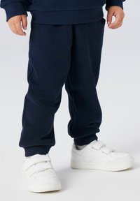 Marineblå joggebukser laget av mykt bomullsmateriale med ribbestrikket midje, kombinert med hvite sneakers med borrelås.