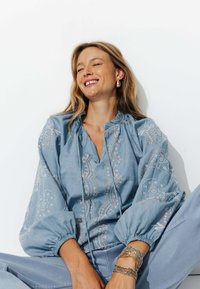 Blouse brodée bleu clair avec une coupe décontractée, des manches ballon, une petite ouverture au niveau du cou et des motifs de broderie pastel décoratifs.