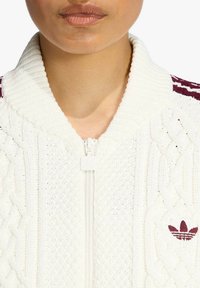 Giacca bianca lavorata a maglia con chiusura a zip e motivo a treccia testurizzato, con accenti color bordeaux inclusi un piccolo logo sul petto.