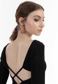 Schwarzes Langarmkleid mit tiefem V-Ausschnitt hinten und überkreuzten Trägern. Das Modell trägt große, metallische Kreiseohrringe, die ihr Profil betonen.