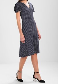 Náměstí modré šaty s květinovým vzorem, s výstřihem ve tvaru V a krátkými rozšířenými rukávy. Spárováno s černými podpatky s tečkovaným vzorem na širokém podpatku.