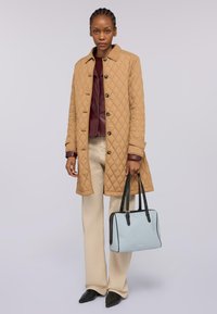 Cappotto beige trapuntato, bottoni scuri, top borgogna a strati, pantaloni crema, scarpe nere, tiene in mano una borsa azzurro chiaro con dettagli neri.