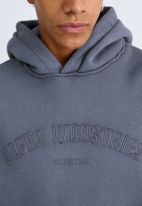 Haut à capuche gris avec poche kangourou, poignets côtelés et texte en relief "ALPHA INDUSTRIES EST. SINCE 1959" à l'avant. Texture de tissu douce.