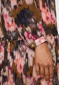 Pitkähihainen mekko, jossa on vaaleanpunaisen, ruskean ja tummansinisen sävyjä. Kankaassa on pehmeä, kuvioitu pinta, ja hihansuissa on napit koristeena.