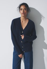 Granatowy kardigan dziany z dekoltem w serek, długimi rękawami i pięcioma kremowymi guzikami z przodu. Miękka faktura i luźny krój.