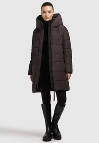 Mørkebrun pufferjakke med hætte, med en quiltet design, lynlås foran og snore detaljer. Modellen har sorte leggings og støvler til knæene på.