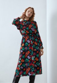 Blomsterkjole med lange ermer, svart bunn, livlige røde, grønne, lilla og hvite mønstre. Avsluttet med et belte i livet.