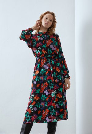 Kukallinen mekko pitkillä hihoilla, musta pohja, kirkkaat punaiset, vihreät, purppurat ja valkoiset kuviot. Viimeistelty vyötärölle solmittavalla vyöllä.