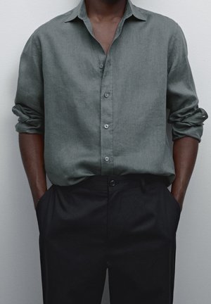 Homme à la peau foncée portant une chemise verte à manches longues boutonnée avec les manches retroussées et un pantalon noir, les mains dans les poches.