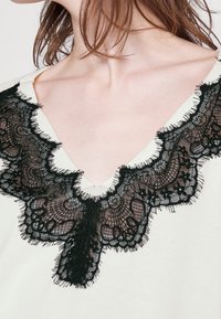 Weißes Kleidungsstück mit V-Ausschnitt, das mit aufwendigen schwarzen Spitzeverzierungen ausgestattet ist und feine Muster sowie eine unregelmäßige Kante für zusätzliche Textur zeigt.