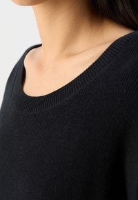 Gros plan d'une personne portant un pull en maille noire avec un col arrondi, montrant le cou et le haut de la poitrine.