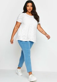 Top blanco de mangas cortas con detalles de ojales, combinado con jeans ajustados azul claro y zapatillas blancas, creando un conjunto casual.